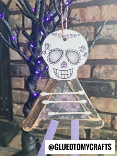 A handmade Day of the Dead craft stick skeleton ornament with a decorated skull and popsicle stick body hangs in front of a black tree with purple lights.