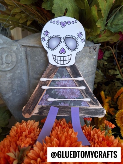 A craft stick skeleton with a triangle body, purple legs, and a decorated skull head is displayed among orange flowers. @GLUEDTOMYCRAFTS is written at the bottom.