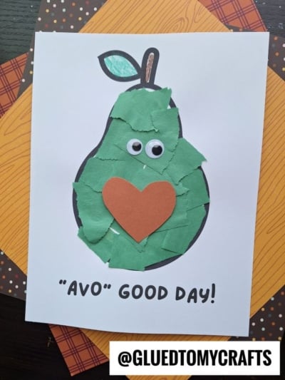 A handmade card featuring a cheerful pear crafted from green paper scraps, googly eyes, and a heart shape—just like a playful paper avocado craft. The text reads AVO GOOD DAY! and @GLUEDTOMYCRAFTS appears at the bottom.