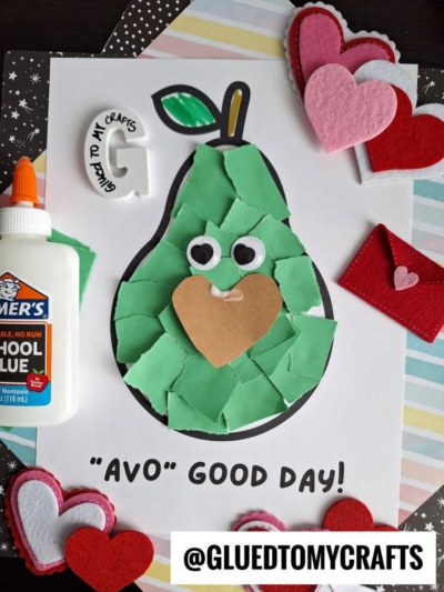 A handmade paper avocado craft features a smiling torn paper avocado with googly eyes and a heart-shaped pit, surrounded by felt hearts, glue, and an "I love crafts" magnet on a striped background. Text says AVO GOOD DAY!.