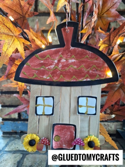 A fall-themed craft of a popsicle stick acorn fairy house, with a red roof, yellow window panes, sunflowers, and mushroom decorations, is displayed in front of autumn leaves.