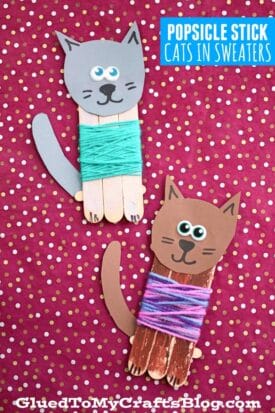 Two popsicle stick cat crafts on a red polka dot background. Each popsicle stick cat has googly eyes, paper ears and tails, drawn-on faces, and colorful yarn sweaters. Text reads “Popsicle Stick Cats in Sweaters.”.