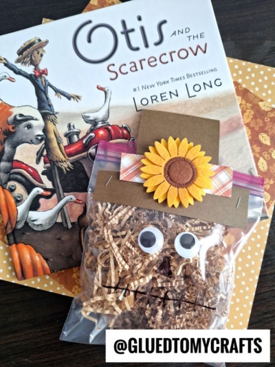 A crinkle paper scarecrow craft bag with googly eyes, a sunflower decoration, and shredded paper sits on top of the book Otis and the Scarecrow by Loren Long. @GLUEDTOMYCRAFTS is written at the bottom.