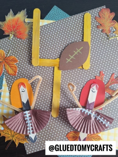 Two handmade cheerleader dolls, including a popsicle stick cheerleader, stand by a craft football goalpost and football, with autumn leaves and polka-dotted paper backgrounds. @GLUEDTOMYCRAFTS is labeled at the bottom.