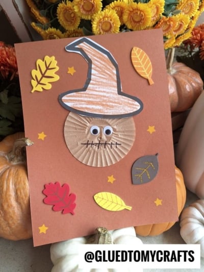 A handmade fall-themed card features a witch hat, googly eyes, and a cupcake liner scarecrow face, decorated with colorful paper leaves and stars, placed among pumpkins. The handle @GLUEDTOMYCRAFTS is at the bottom.