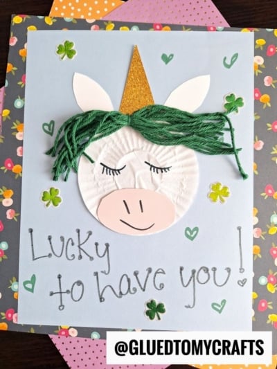 Handmade cupcake liner unicorn card with a paper face, gold horn, green yarn hair, and drawn features, decorated with green clover stickers and the message Lucky to have you! on a blue background.