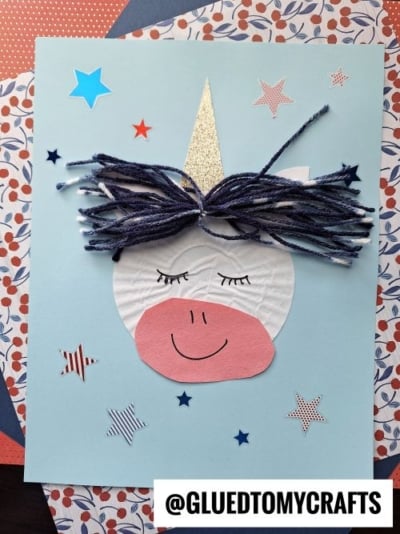 A handmade cupcake liner unicorn card features a white and pink unicorn face with closed eyes and a yarn mane on blue paper, adorned with star stickers. The handle @GLUEDTOMYCRAFTS appears at the bottom.