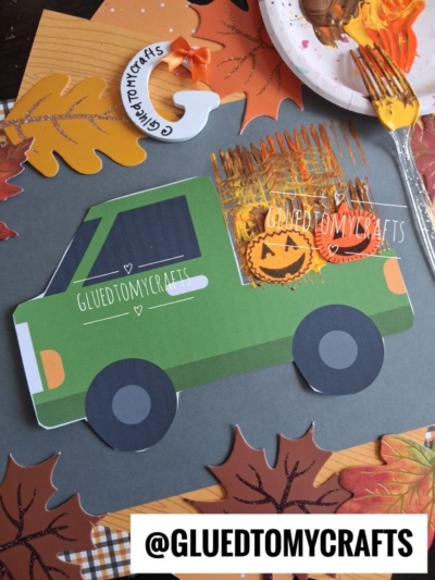 Paper craft of a green paper farm hay truck carrying painted pumpkins on gray paper, surrounded by cutout leaves and crafting supplies. @GLUEDTOMYCRAFTS is displayed on the truck and at the bottom of the image.