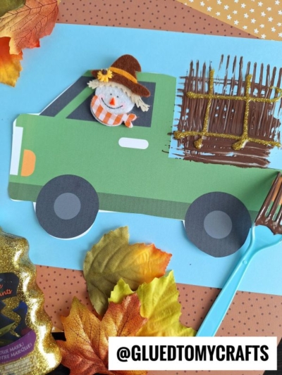A green paper hay truck with a smiling scarecrow in the window carries a textured brown load, fork painted to look like hay. Autumn leaves and craft supplies surround the truck on a blue background. @GLUEDTOMYCRAFTS is at the bottom.