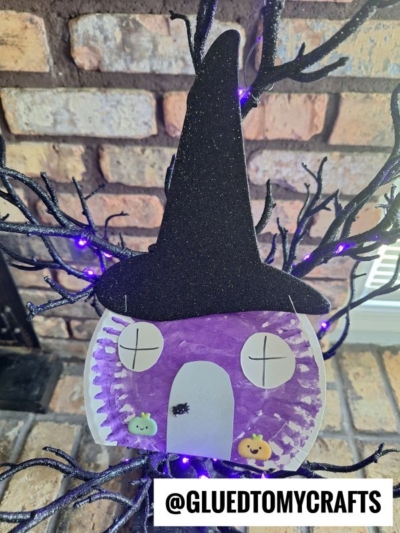 A Halloween witch house paper plate craft featuring a purple face with a black hat, paper eyes, and door, adorned with stickers. It hangs on black branches with purple lights. @GLUEDTOMYCRAFTS text is at the bottom.