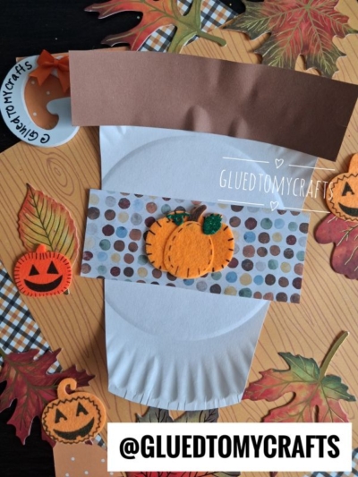 A coffee cup-shaped paper craft decorated with a felt pumpkin, surrounded by autumn-themed paper cutouts, leaves, and jack-o-lanterns on a wooden surface evokes cozy paper plate creations and the inviting warmth of pumpkin spice.