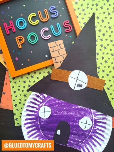 A whimsical paper craft witch face with a purple crayon mask and black hat is displayed next to a sign spelling Hocus Pocus in colorful letters on star-patterned paper, making it the perfect companion to any witch house paper plate craft.