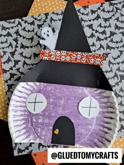 A Halloween witch house paper plate craft featuring a witch’s face made from a purple plate, with white X eyes, a black hat, an orange band with skulls, a small ghost decoration, and Halloween-themed paper in the background.