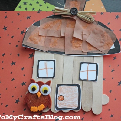 A popsicle stick acorn fairy house with a brown paper roof, felt owl, orange windows, and a button door sits on an orange background with black stars.