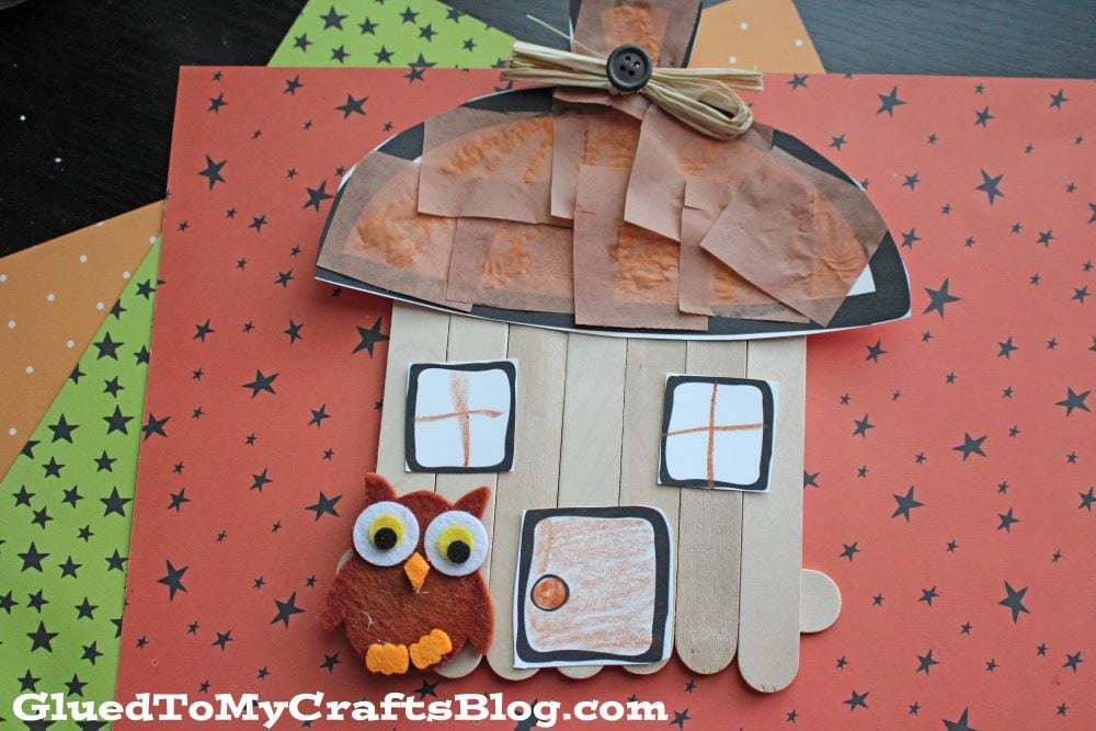 A popsicle stick acorn fairy house with a brown paper roof, felt owl, orange windows, and a button door sits on an orange background with black stars.