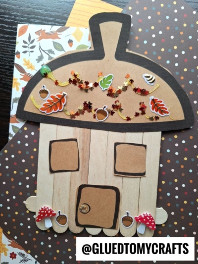 A fall-themed popsicle stick acorn fairy house crafted from popsicle sticks and shaped like an acorn, decorated with autumn stickers, sequins, leaves, and small mushroom cutouts. The username @GLUETOMYCRAFTS is displayed.