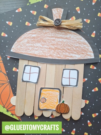 A fall-themed popsicle stick acorn fairy house craft hut made from popsicle sticks, colored paper, and raffia, set on Halloween-patterned paper with candy corn designs. @GLUEDTOMYCRAFTS is written in a green banner.