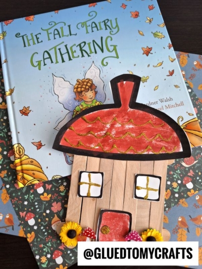 A children's book titled The Fall Fairy Gathering lies on patterned paper with a popsicle stick acorn fairy house craft, decorated with flowers, mushrooms, and glitter. @GLUEDTOMYCRAFTS is written at the bottom.