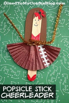 A popsicle stick cheerleader craft features a paper skirt, brown hair, a red bow, gold pipe cleaner pom-poms, and a football field background. Text reads “POPSICLE STICK CHEERLEADER.”.