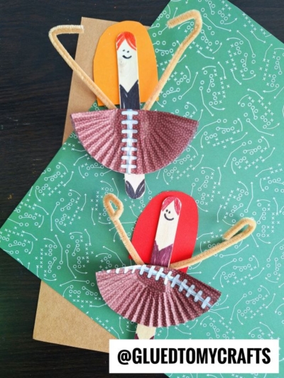 Two handmade ballerina crafts crafted with orange and brown paper, cupcake liners for skirts, pipe cleaners for arms, and smiling faces drawn on wooden clothespins. Inspires creative play—pair with a popsicle stick cheerleader for extra fun! Background features patterned paper.