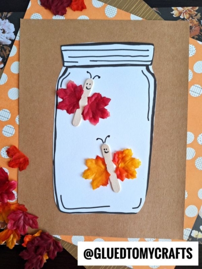 A paper craft features a mason jar outline with two leaf butterflies made from craft sticks and colorful fabric leaves, each with a smiling face. The background includes orange patterned paper and loose fabric leaves.