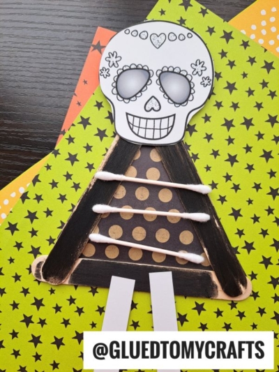 A Day of the Dead craft featuring a popsicle stick skeleton skull with decorative designs on a triangle made from black-painted sticks, polka-dot paper, and cotton swabs, set against a star-patterned background. @GLUEDTOMYCRAFTS.