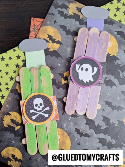 Two Halloween-themed bookmarks made from colored popsicle sticks—one green with a skull and crossbones, the other purple with a ghost. They rest on black paper with bat and moon patterns. Text: @GLUEDTOMYCRAFTS. Perfect for a witch's potion-themed craft!.