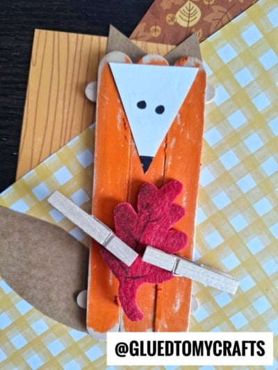 A leaf holding fox craft made from orange popsicle sticks, with a white triangular face, black eyes and nose, brown paper tail, clutching a red felt leaf and clothespin, displayed on patterned paper. @GLUEDTOMYCRAFTS is printed below.
