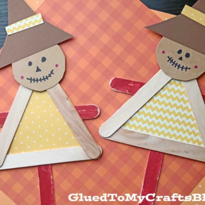 Two handmade popsicle stick scarecrow puppets made from popsicle sticks, paper, and markers are displayed on orange plaid paper. Each scarecrow has a brown paper hat, a yellow triangular body, and a smiling face.