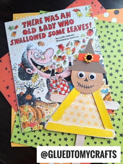 A children’s book titled “There Was an Old Lady Who Swallowed Some Leaves!” rests on colorful polka dot paper, next to a handmade popsicle stick scarecrow puppet with a witch hat and yellow dress. @GLUEDTOMYCRAFTS is labeled below.