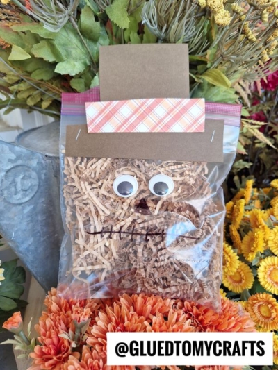 A clear plastic bag filled with brown crinkle paper is decorated as a paper scarecrow craft, complete with googly eyes, a stitched mouth, and a paper hat. Orange and yellow flowers surround it, with @gluedtomycrafts labeled at the bottom.