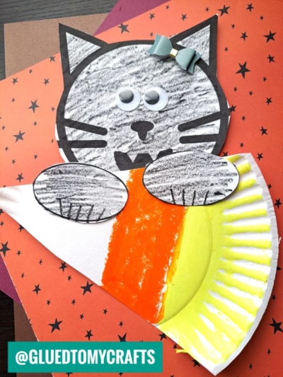 A paper craft of a black and white cat with googly eyes and a blue bow, holding a paper plate candy corn, set on a star-patterned orange background.