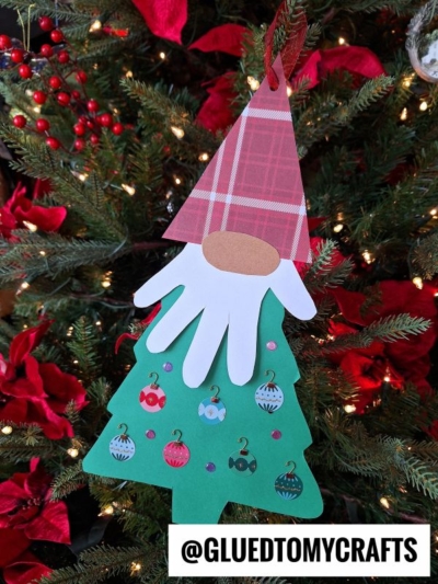 A handmade christmas tree gnome ornament hangs on a tree. The gnome has a red plaid hat, a white paper handprint beard, a brown oval nose, and a green tree-shaped body decorated with ornaments.