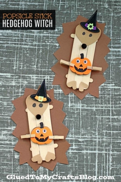 Two popsicle stick hedgehog witches made from brown paper and decorated with charming hats and mini pumpkins are displayed on a textured gray background, with “Popsicle Stick Hedgehog Witch” text at the top.