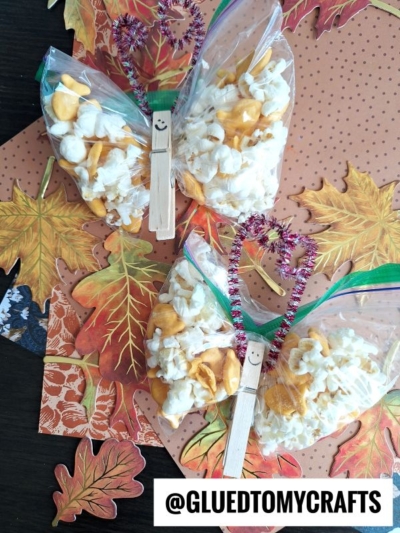 Two fall butterfly snack bags clipped with clothespins, decorated as butterflies, are filled with popcorn and goldfish crackers. Each butterfly features pipe cleaner antennae, a smiley face, and sits on autumn leaf-patterned paper.