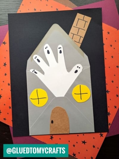 A Halloween craft showing a handprint haunted house made from paper, with a white handprint ghost floating at the front, yellow windows, and a brown door. The background has orange and black star-patterned paper.