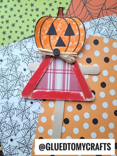 A jack-o-lantern scarecrow with a pumpkin head, made from craft sticks and plaid paper, sits on Halloween-themed paper adorned with spiderwebs, stars, and dots.