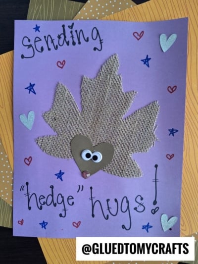 A purple card with a burlap leaf hedgehog, a heart-shaped face with googly eyes, hearts, and text reading “Sending hedge hugs!” The handle @GLUEDTOMYCRAFTS is at the bottom.