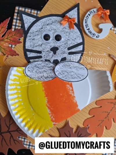 A paper plate candy corn cat craft with orange, yellow, and white sections, surrounded by paper leaves on a wood-patterned background. The cat has a gray face, googly eyes, and small orange bows.