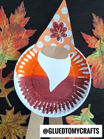 A paper plate fall gnome craft painted in vibrant autumn colors features a whimsical white beard and a bright orange hat, surrounded by artificial autumn leaves. The image includes the text @GLUEDTOMYCRAFTS at the bottom.
