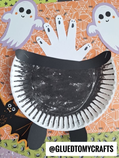 Create a spooky Paper Plate Witch Cauldron craft this Halloween, featuring a paper plate cauldron painted black, a ghostly hand cutout, and bat and ghost decorations—all set against an orange patterned background.