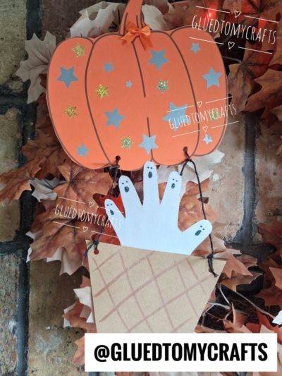 A jack-o-lantern hot air balloon craft with silver stars floats above a paper handprint ghost in a brown basket, decorated with fall leaves. Text reads @GLUEDTOMYCRAFTS.