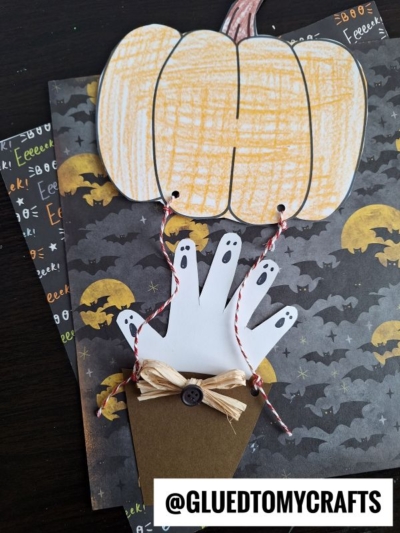 A Halloween-themed craft featuring a jack-o-lantern hot air balloon with three ghostly white handprint passengers, a brown paper basket with a bow, and a bat and moon background. @GLUEDTOMYCRAFTS is written at the bottom.