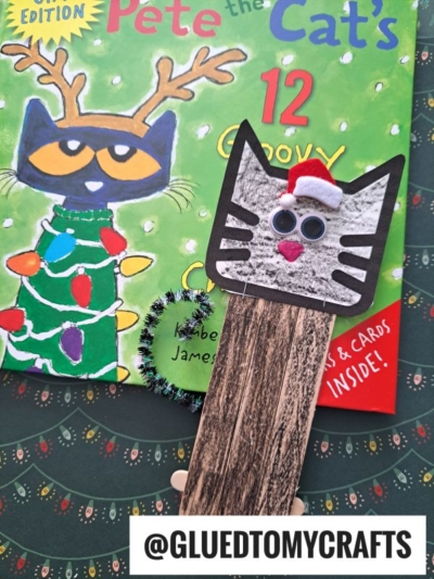 A handmade popsicle stick cat bookmark with a Santa hat and googly eyes rests on the festive cover of Pete the Cat’s 12 Groovy Days of Christmas book. The username @GLUEDTOMYCRAFTS appears at the bottom.