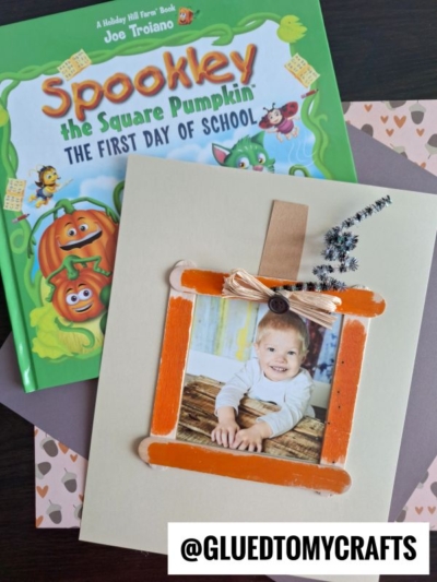 A children’s book titled Spookley the Square Pumpkin and a pumpkin-themed photo frame craft featuring a child’s picture. Craft materials include orange sticks, decorative paper, ribbon, and a button—perfect for creating your own popsicle stick pumpkin patch frame. @GLUEDTOMYCRAFTS text overlay.