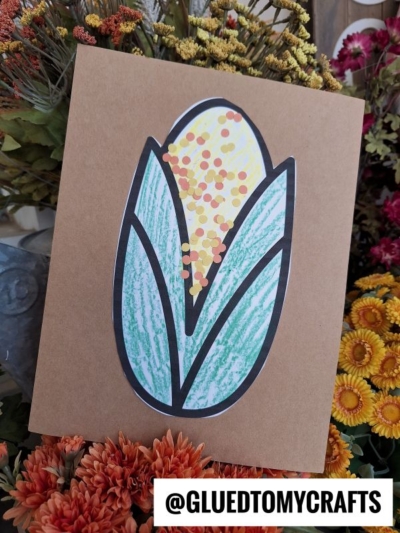 A handmade card with a corn-on-the-cob design, inspired by a classic indian corn paper craft and colored with green leaves topped with yellow and orange dots, is displayed among colorful flowers. The handle @GLUEDTOMYCRAFTS is visible at the bottom.