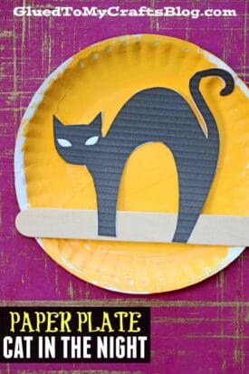 A yellow paper plate features a black cat cutout with arched back and white eyes, above a wooden craft stick, against a purple textured background. Perfect for halloween night crafts, text reads, PAPER PLATE CAT IN THE NIGHT.