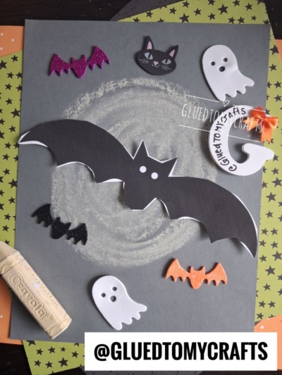 A Halloween craft scene with paper bats, ghosts, and cats on dark paper, chalk swirls, a crayon, and a star-patterned background captures the magic of a chalk art bat in the night. @GLUEDTOMYCRAFTS is printed at the bottom.