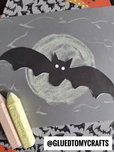 A black paper bat with white eyes is glued to a gray paper with a chalk-drawn moon, creating the look of a chalk art bat in the night. Two Crayola chalk pieces rest nearby. @GLUEDTOMYCRAFTS is written at the bottom.