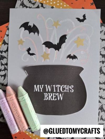 A Halloween-themed chalk art witch cauldron features MY WITCHS BREW text, pastel chalk swirls, black bats, and gold stars on light paper, with colorful Crayola chalks nearby. @GLUEDTOMYCRAFTS watermark is at the bottom.